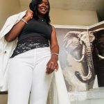 Woman smiling, wearing black top and white pants, standing indoors near a large animal illustration, holding a white jacket over her shoulder.