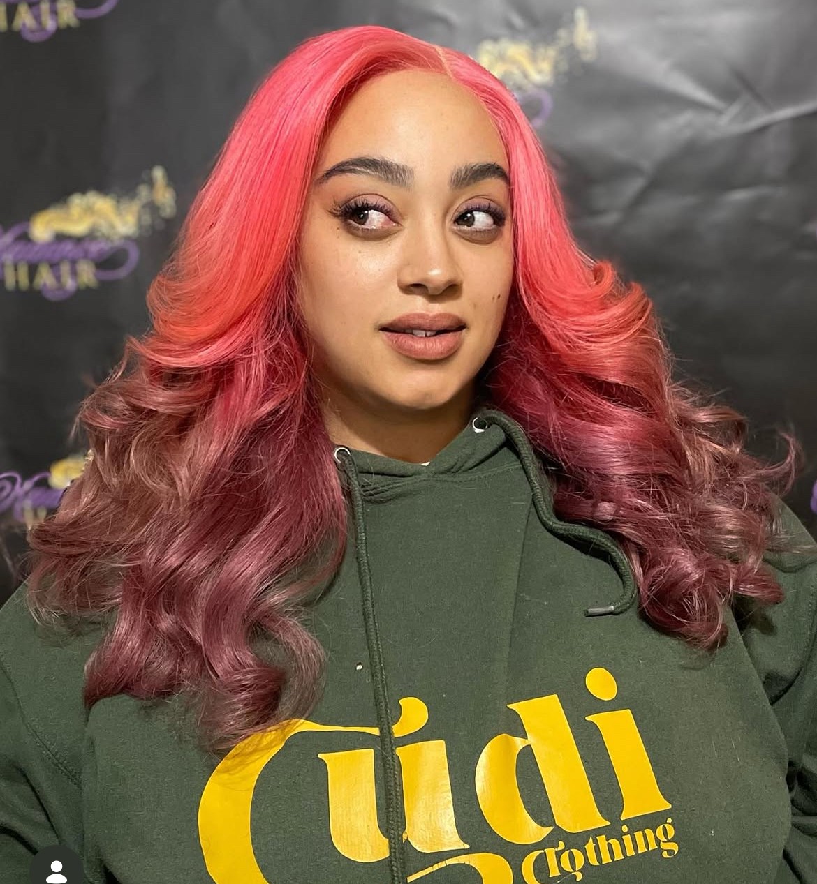 Young woman with pink and purple wavy hair wearing a green hoodie with yellow text, looking to the side.