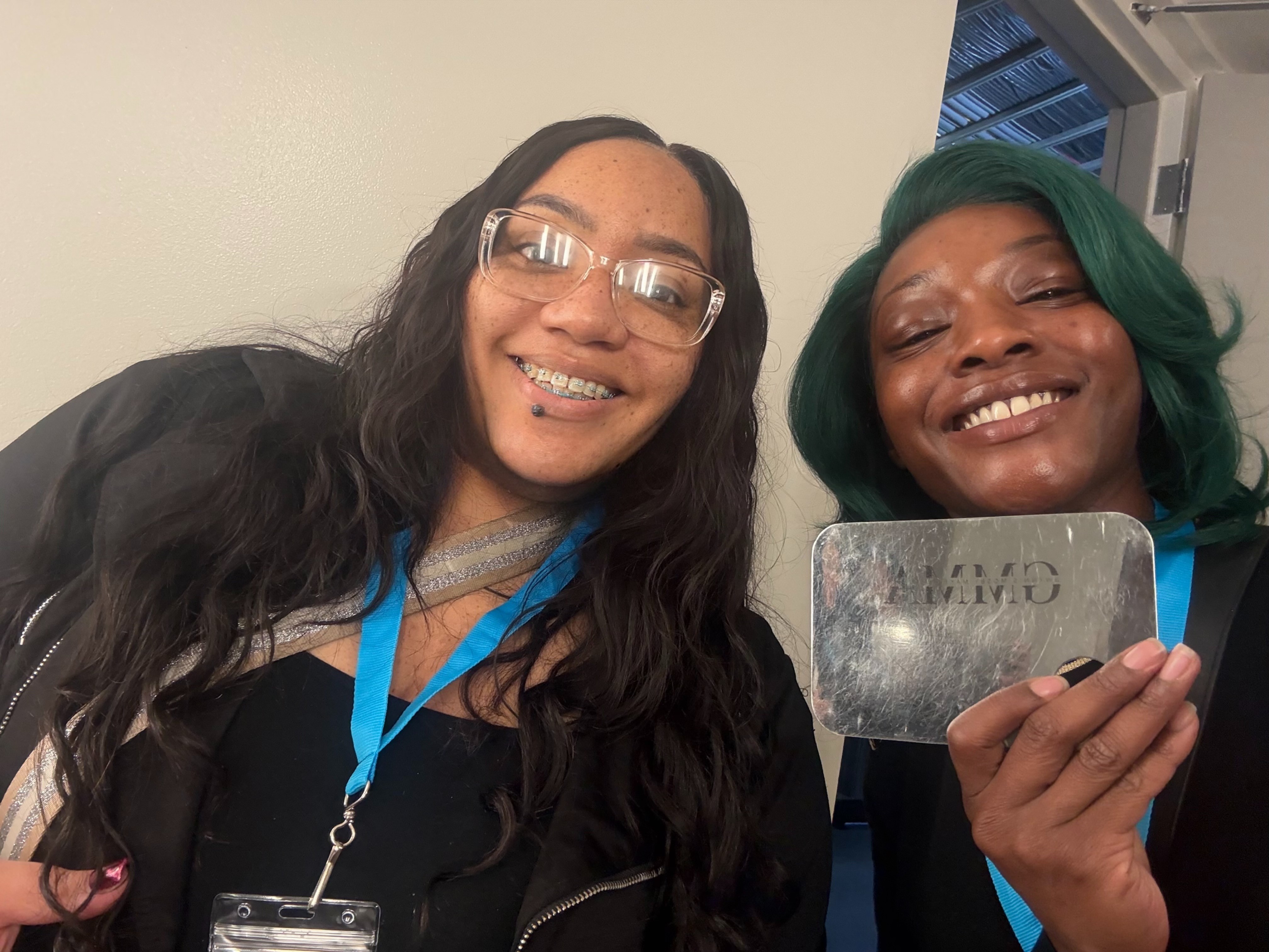 Two women smiling, one holding a silver award or plaque, indoors with a window in the background.