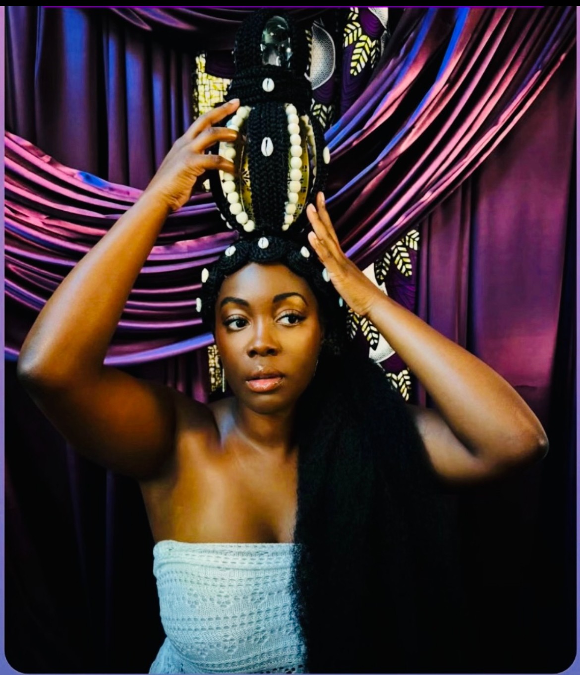 Woman with elaborate headpiece, hands adjusting it, purple and pink draped background, strapless top, serious expression.