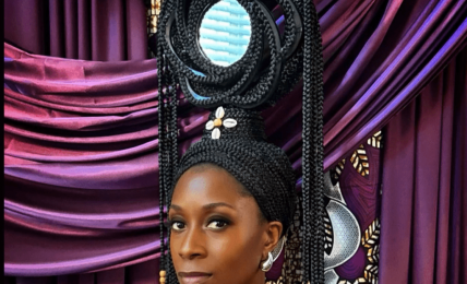 Woman with elaborate hairstyle and jewelry in front of colorful draped fabric backdrop.