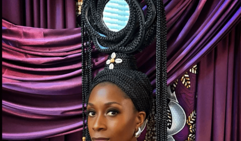 Woman with elaborate hairstyle and jewelry in front of colorful draped fabric backdrop.