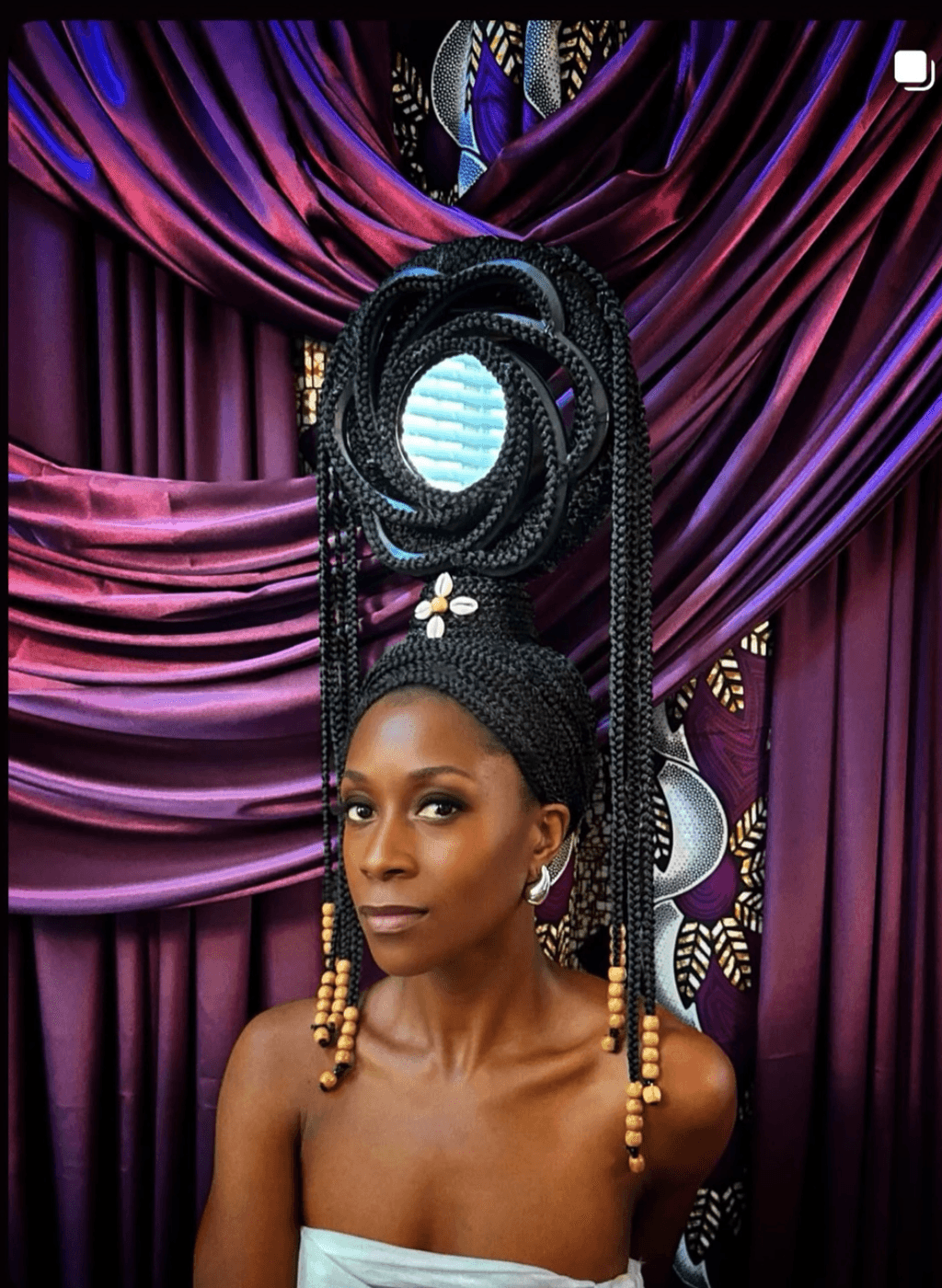 Woman with elaborate hairstyle and jewelry in front of colorful draped fabric backdrop.