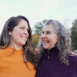 Two women smiling and looking at each other outdoors on a sunny day.