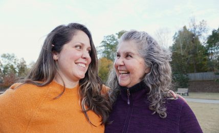 Two women smiling and looking at each other outdoors on a sunny day.