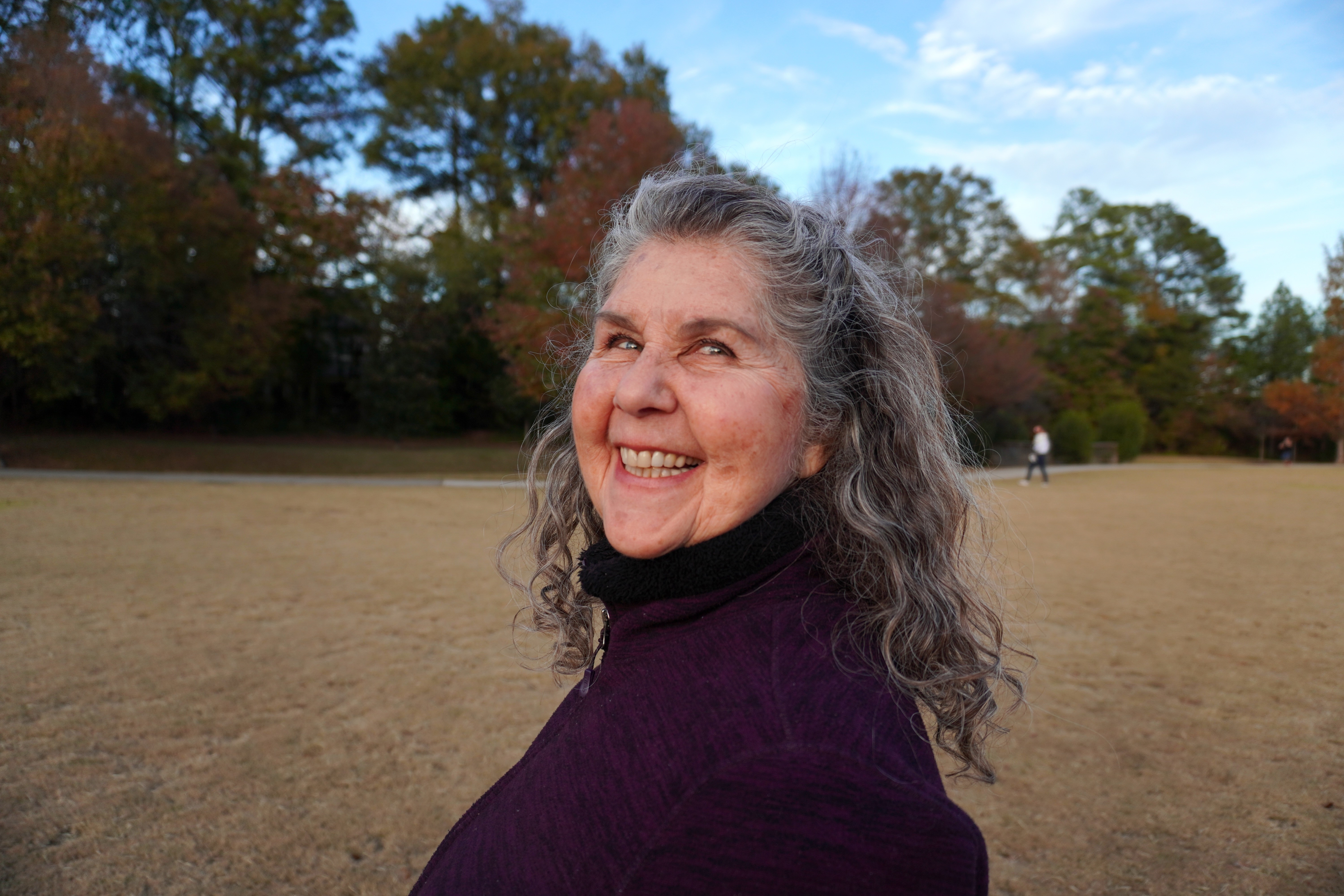 Smiling woman with gray hair outdoors in a park, wearing a dark purple sweater, trees in background.