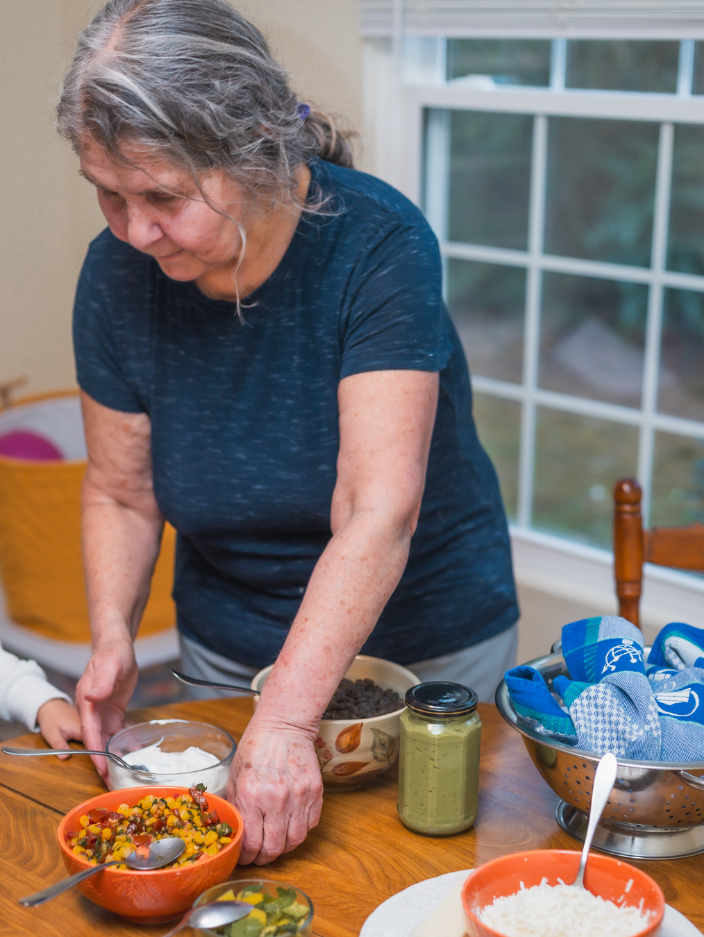 Older woman with gray hair in a blue shirt preparing food at a table near a window, with bowls and jars present.