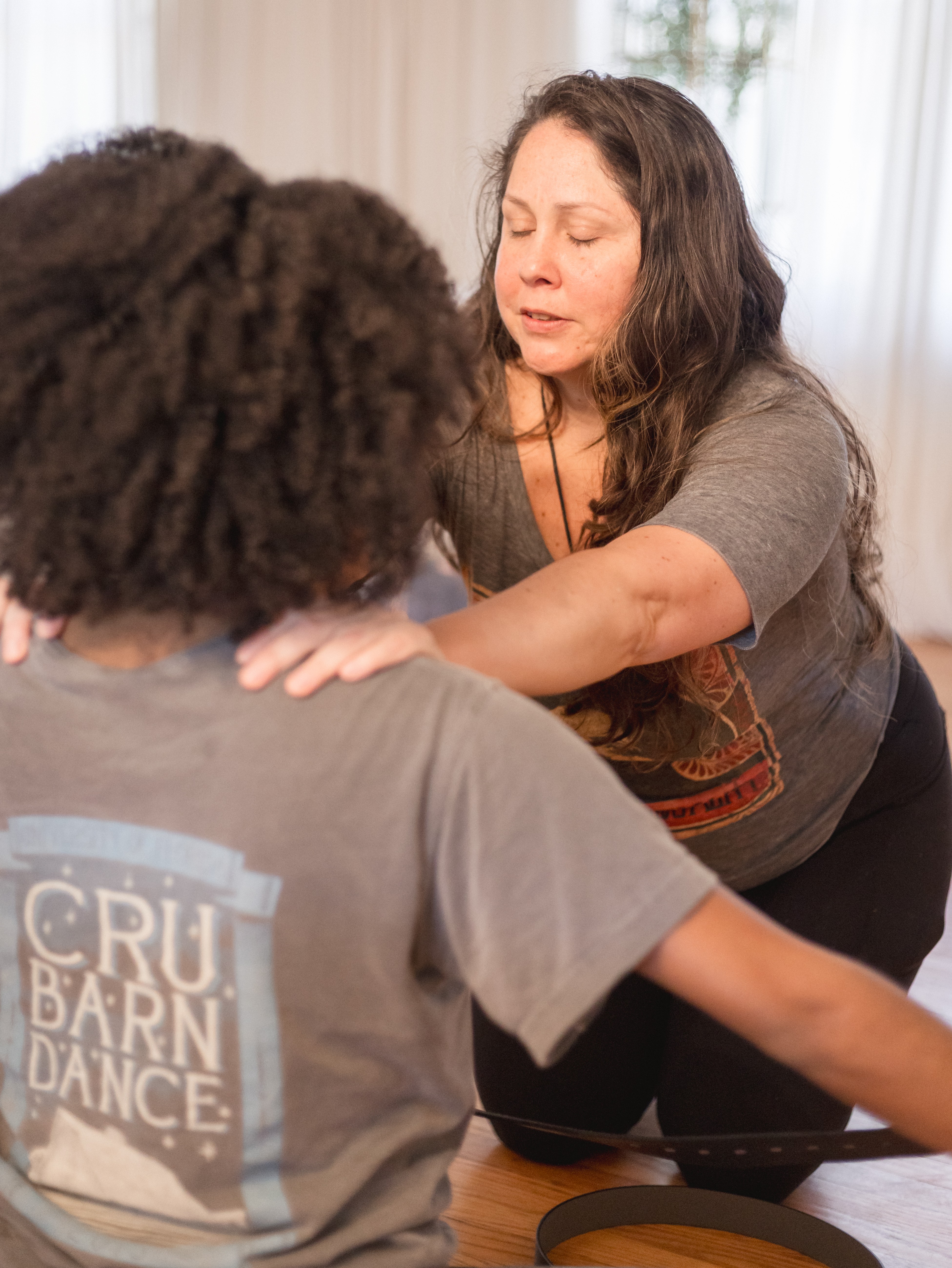 Woman with long dark hair and gray shirt appears to be guiding or assisting a person with curly hair wearing a gray T-shirt, indoors.