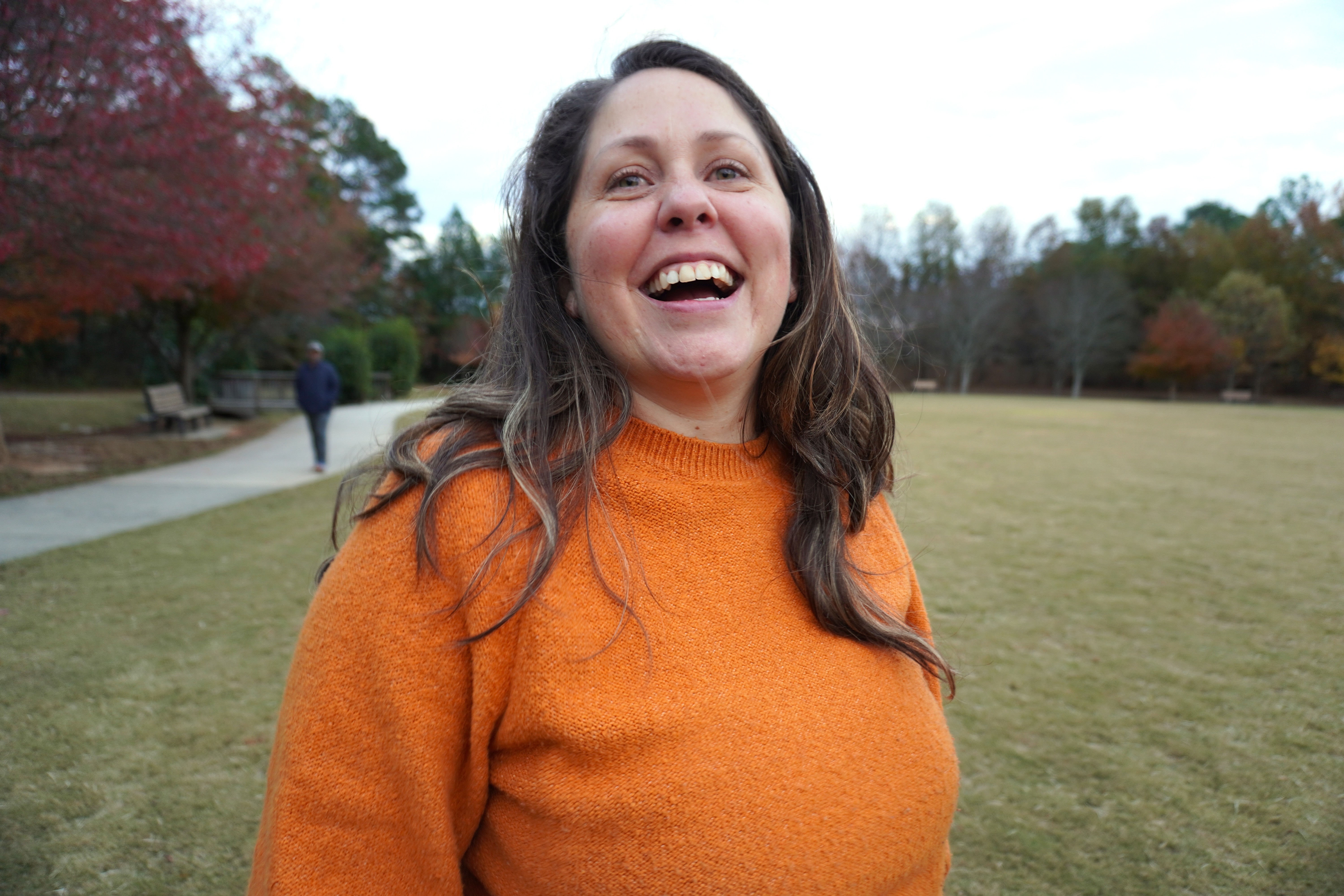 Woman with long hair smiling outdoors in a park, wearing an orange sweater, trees in background.