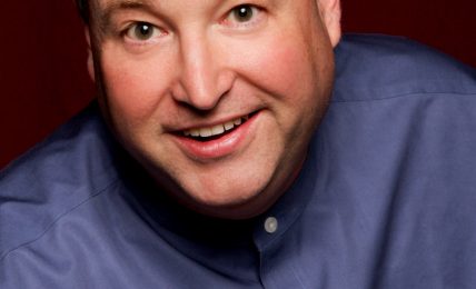 Portrait of a smiling man with short hair, wearing a blue shirt, against a dark red background.