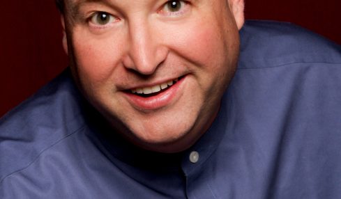 Portrait of a smiling man with short hair, wearing a blue shirt, against a dark red background.