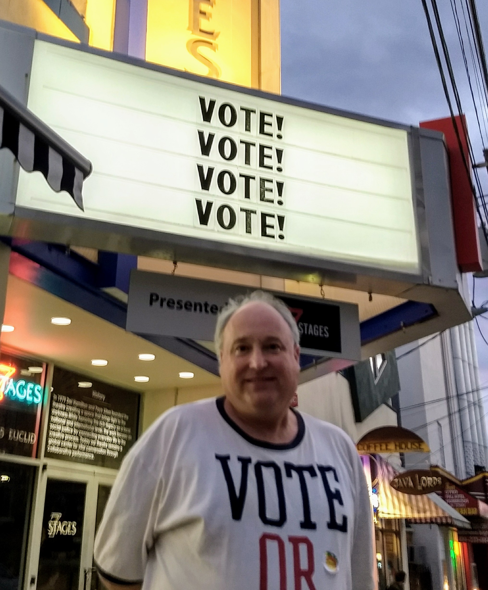 Man wearing a shirt with 'VOTE' standing in front of a voting sign with 'VOTE!' repeated four times.