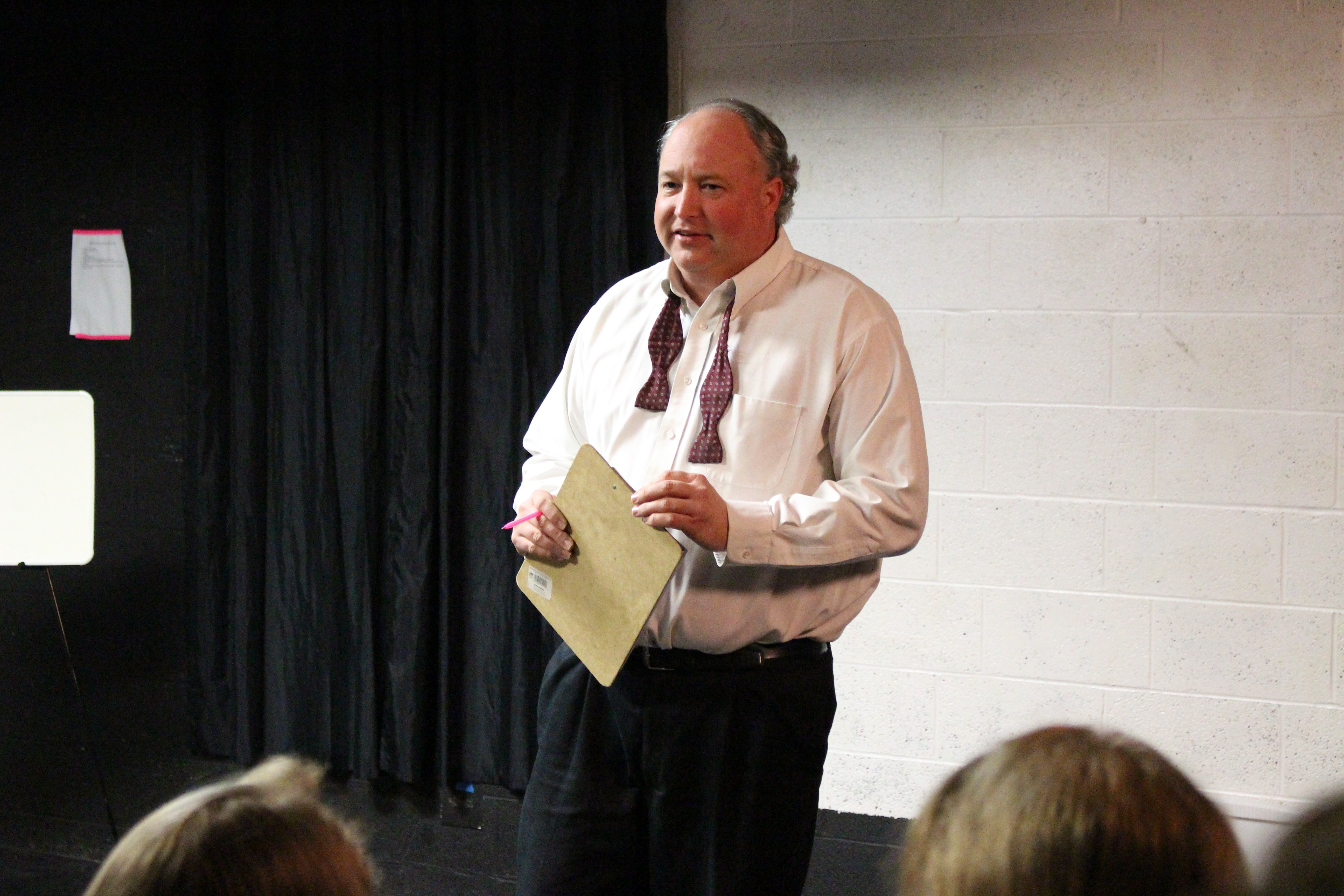 Man standing with a paper and folder, speaking or presenting, in a room with a white wall and black curtain behind him.