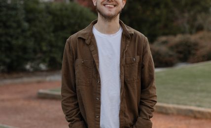 Young man smiling outdoors in a park with trees and bushes in the background.