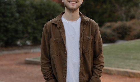 Young man smiling outdoors in a park with trees and bushes in the background.