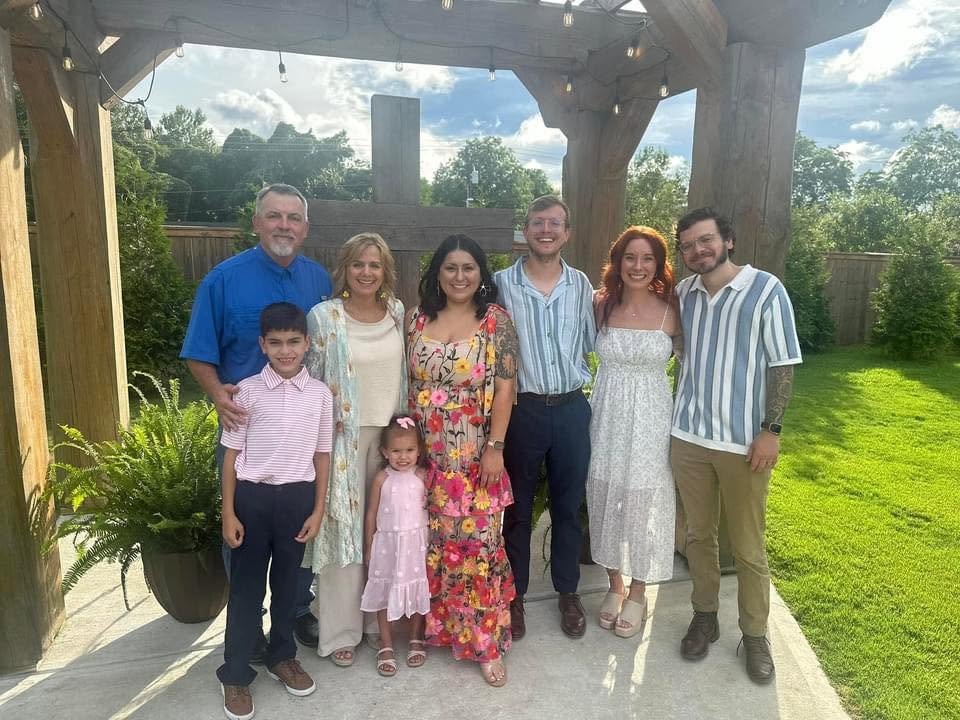 Group of nine people standing outdoors under a wooden structure, smiling, with green grass and trees in background.