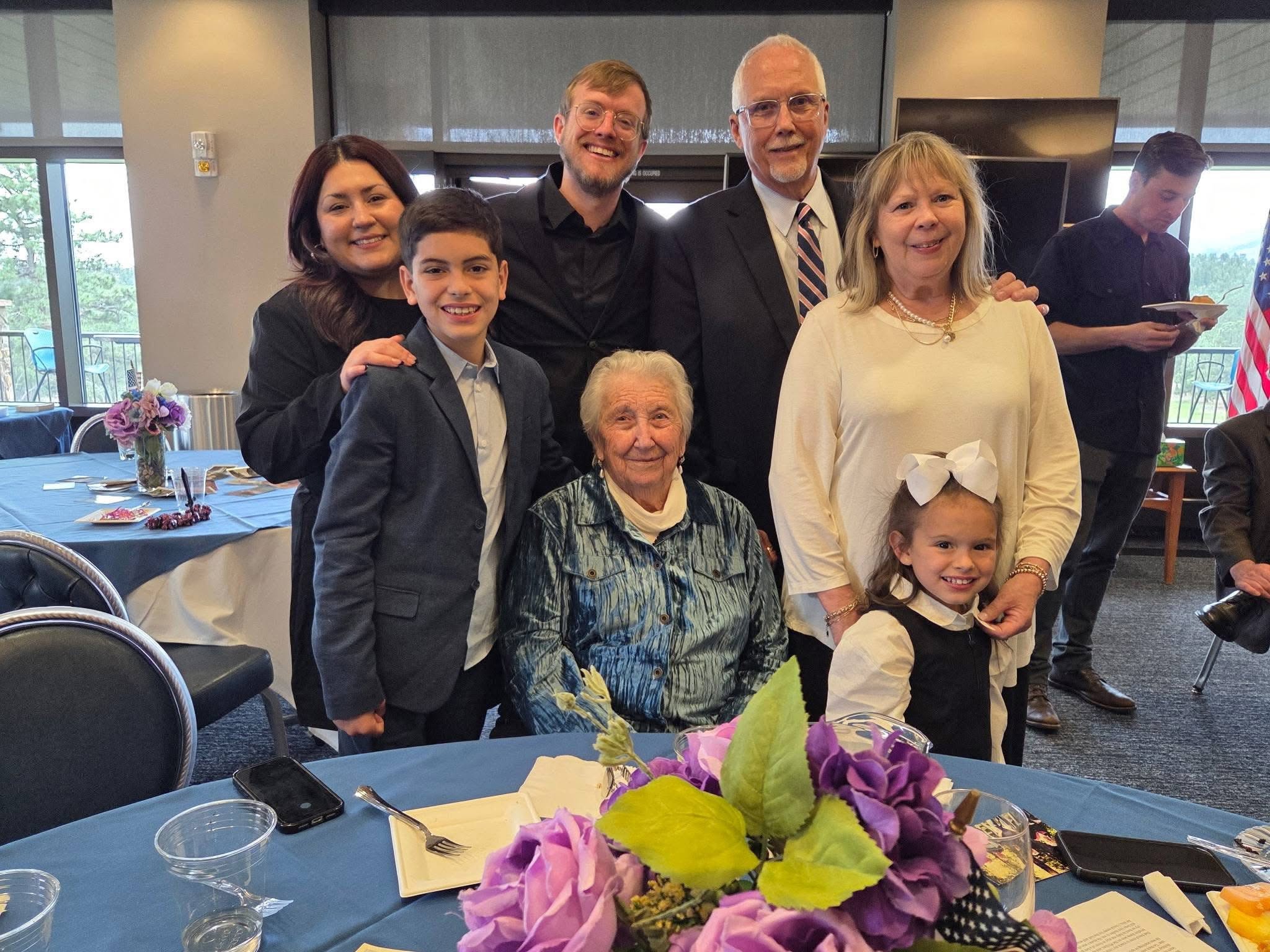 Group of seven people, including children and adults, gathered around a table with a floral centerpiece, smiling indoors.