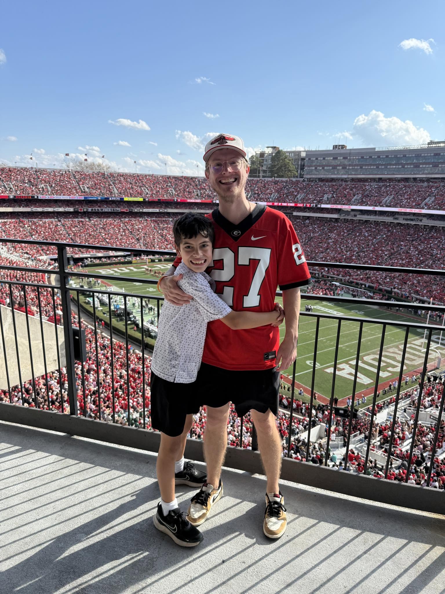Two people, a child and an adult, stand together at a stadium with a football field in the background, smiling.