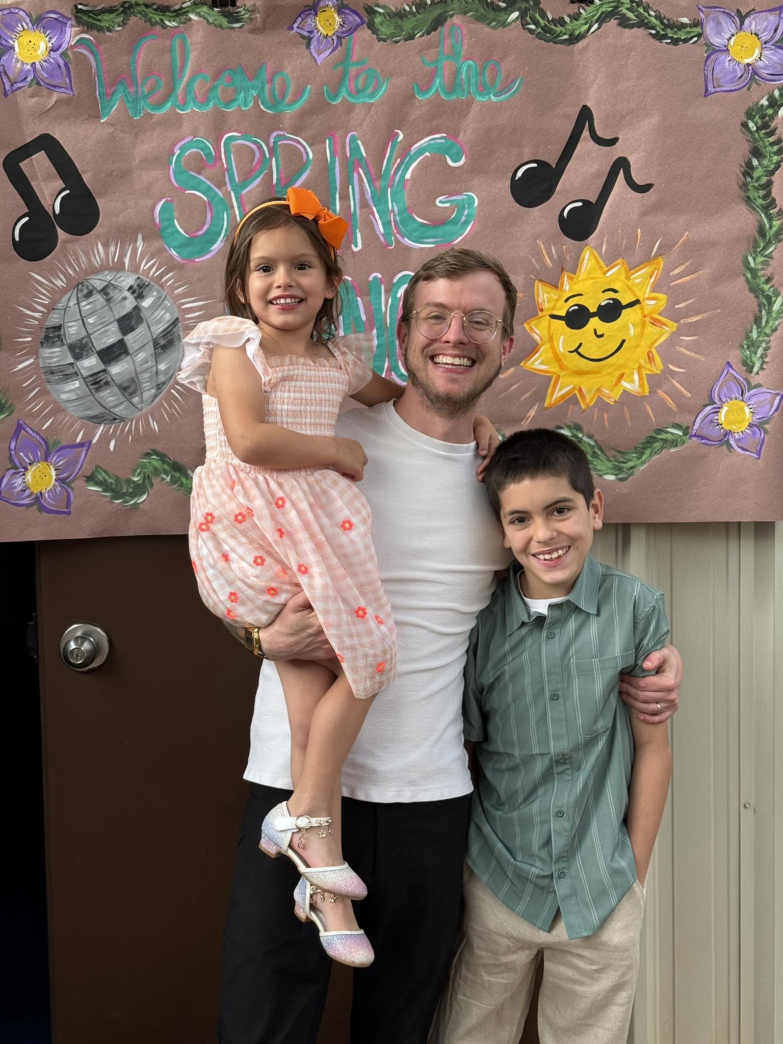 Three people smiling in front of a spring-themed decorated backdrop with musical notes and a sun wearing sunglasses.