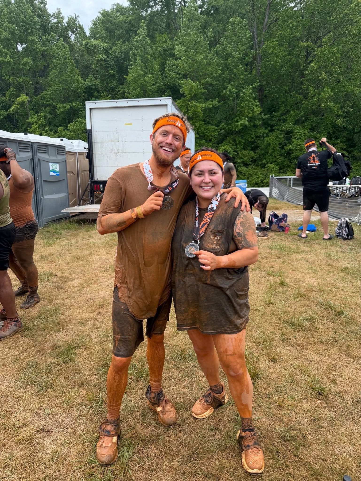 Two smiling people wearing orange headbands and medals, standing outdoors with trees and people in the background.