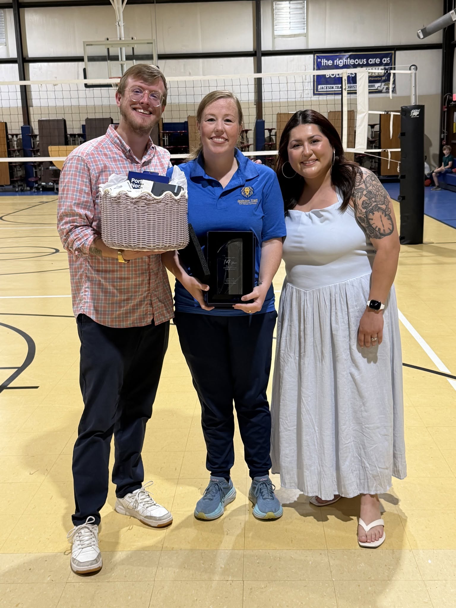 Three people standing in a gymnasium, smiling, with a basket and a trophy, one person in a blue uniform, two in casual clothing.