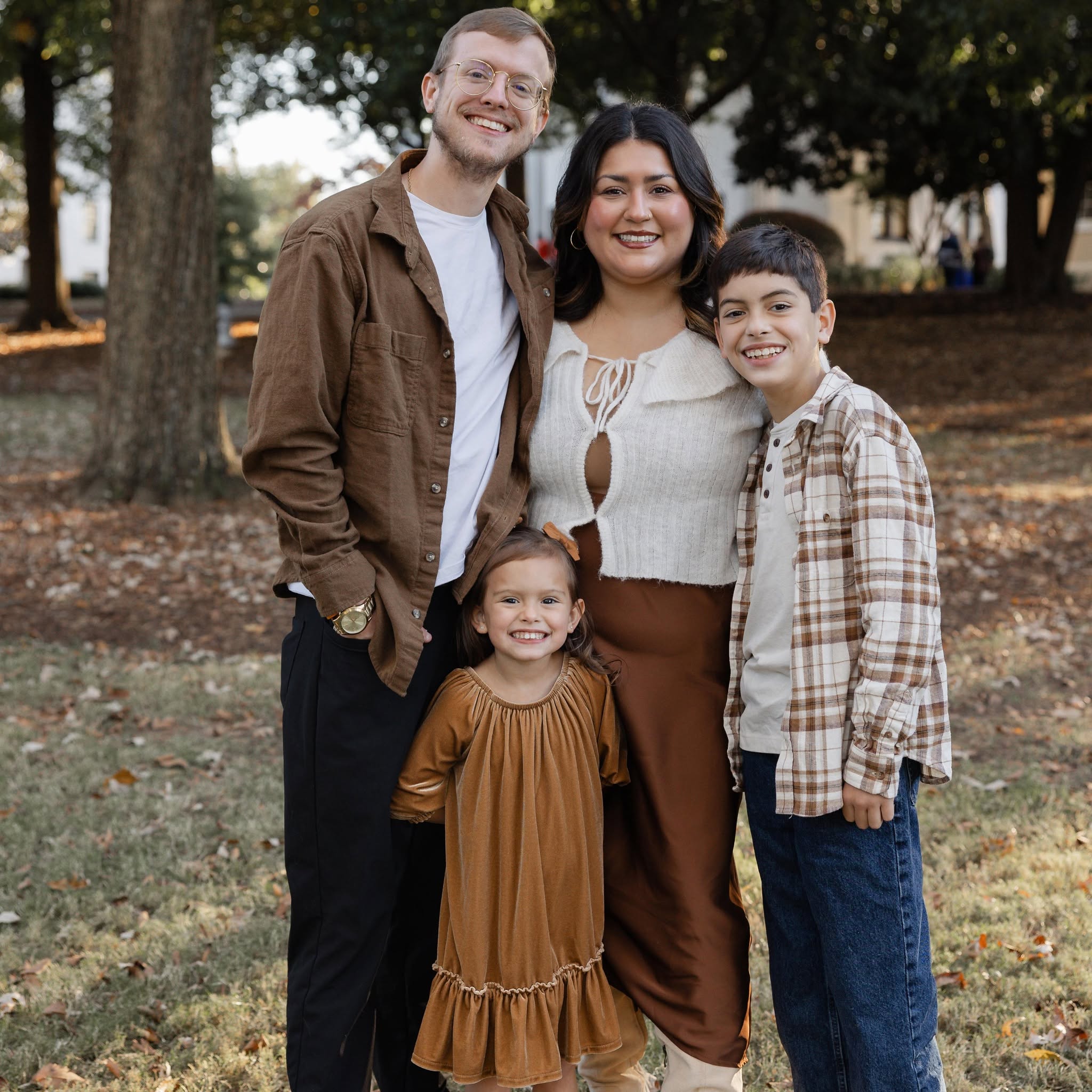 Group of four people smiling outdoors, standing on grass with trees in background.