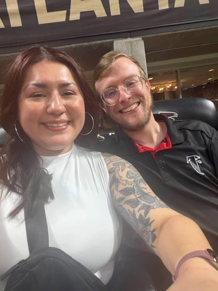 Two smiling people, a woman with dark hair and a man with glasses, sitting close together indoors.
