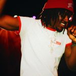 Person with dreadlocks wearing a red beanie and white t-shirt, smiling and dancing in a dark setting.