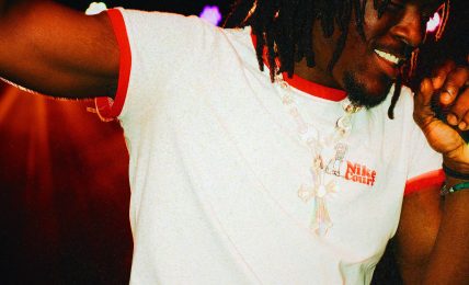 Person with dreadlocks wearing a red beanie and white t-shirt, smiling and dancing in a dark setting.