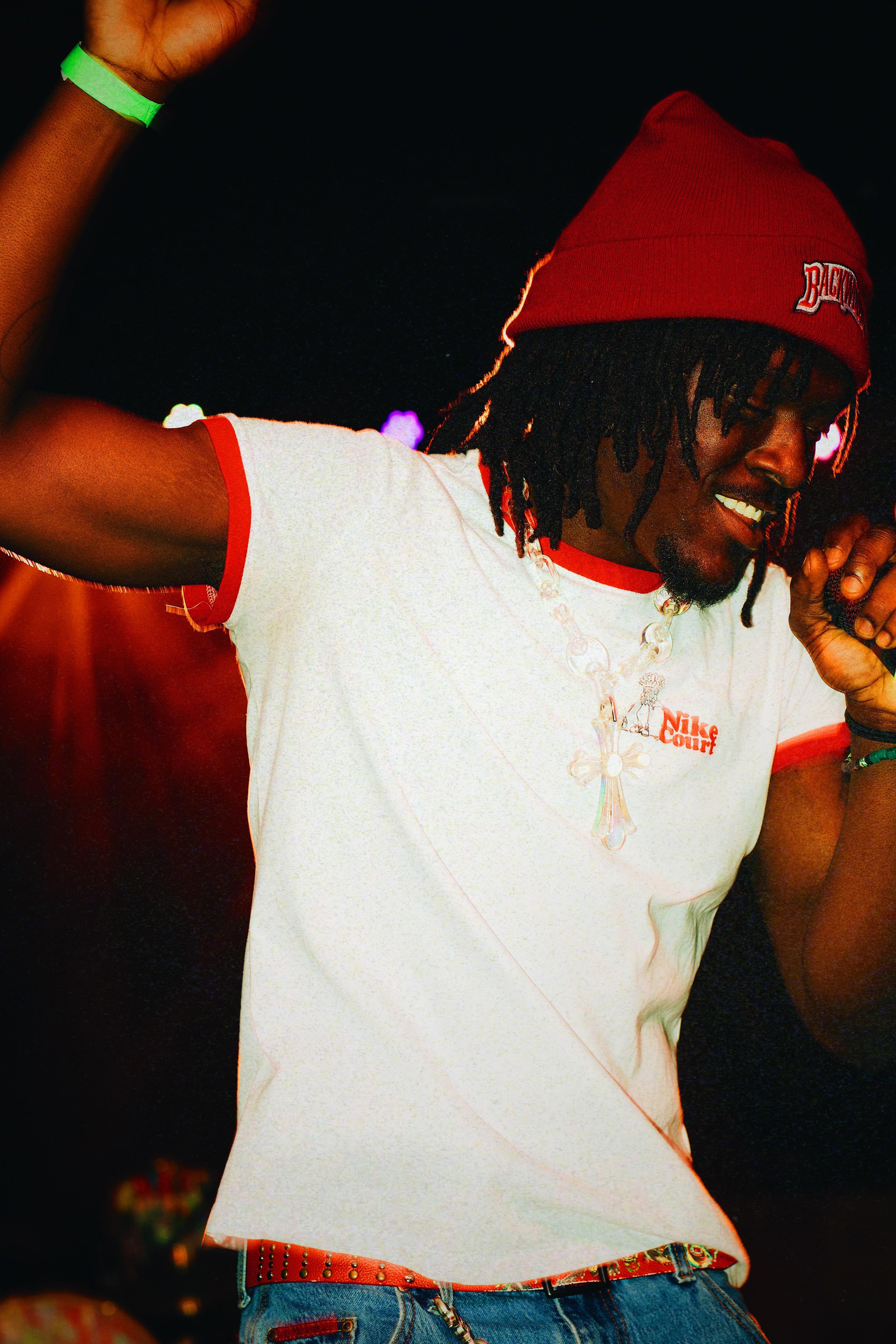 Person with dreadlocks wearing a red beanie and white t-shirt, smiling and dancing in a dark setting.