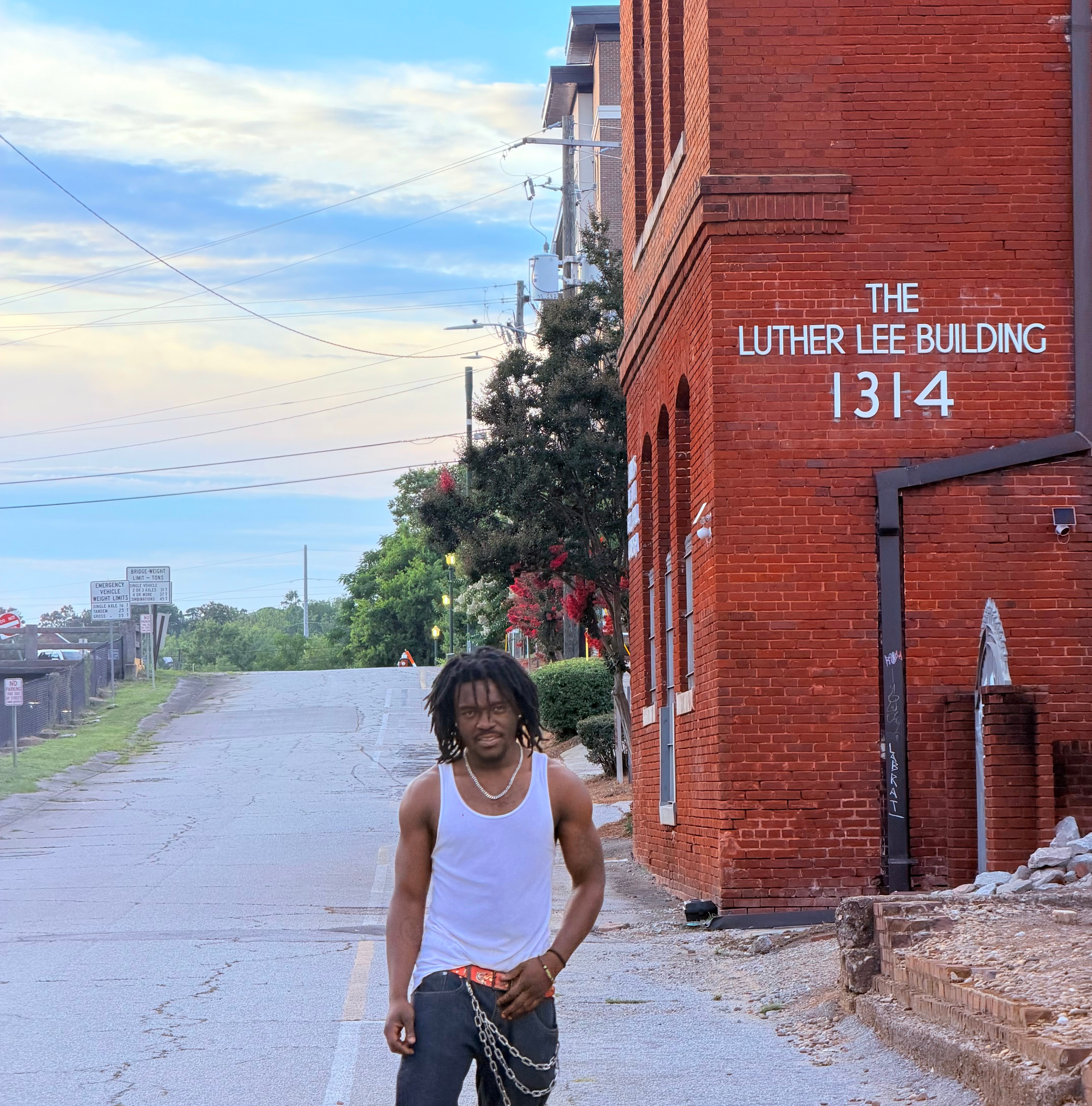 Young man with dreadlocks in a white tank top standing on a street near a red brick building with white text.