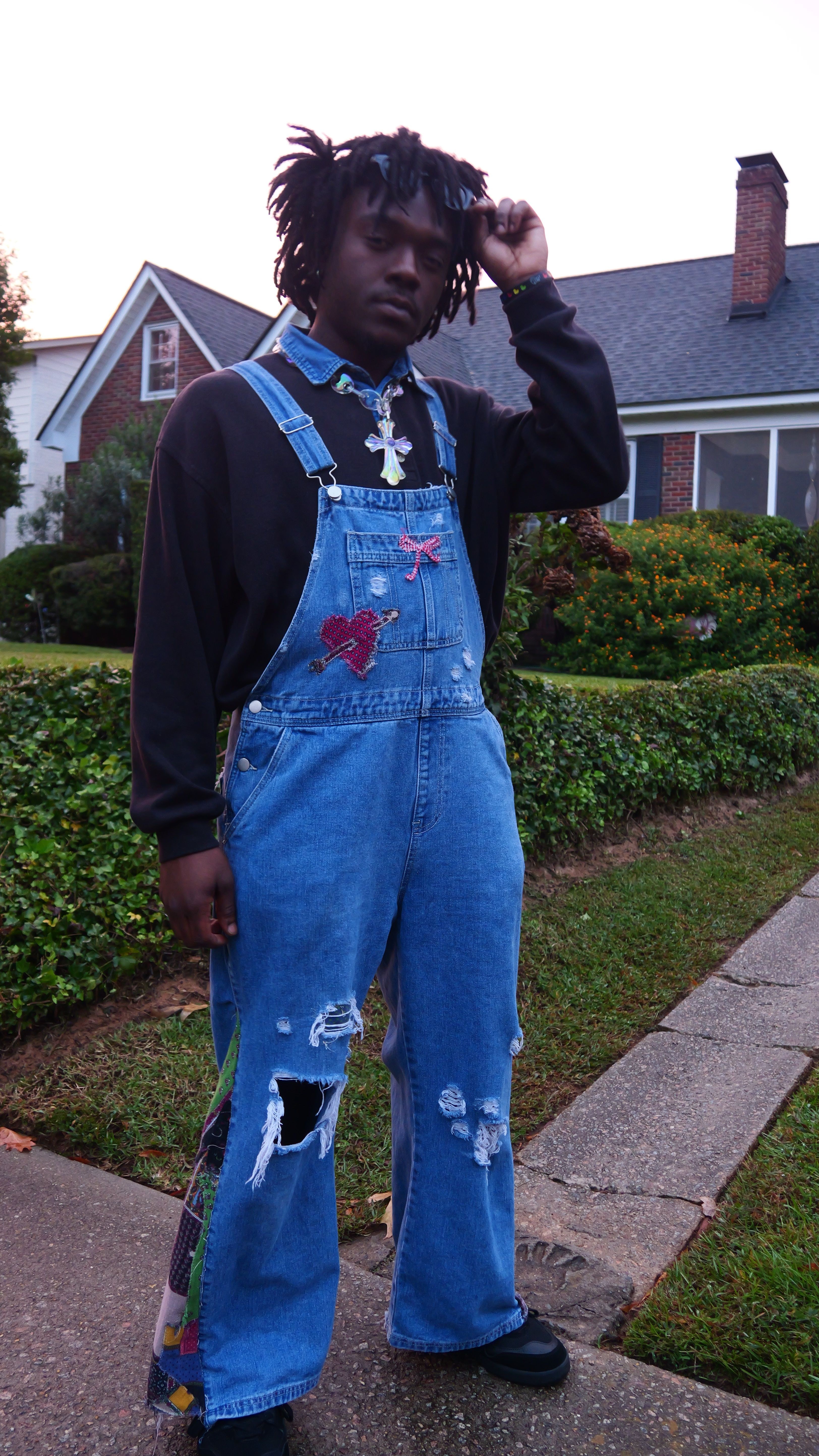 Person standing outdoors on sidewalk, talking on phone, wearing black jacket and blue jeans with holes, in front of houses and greenery.