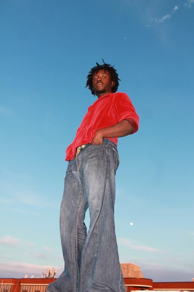 Person standing outdoors against a blue sky, wearing a red shirt and gray pants, with arms crossed.