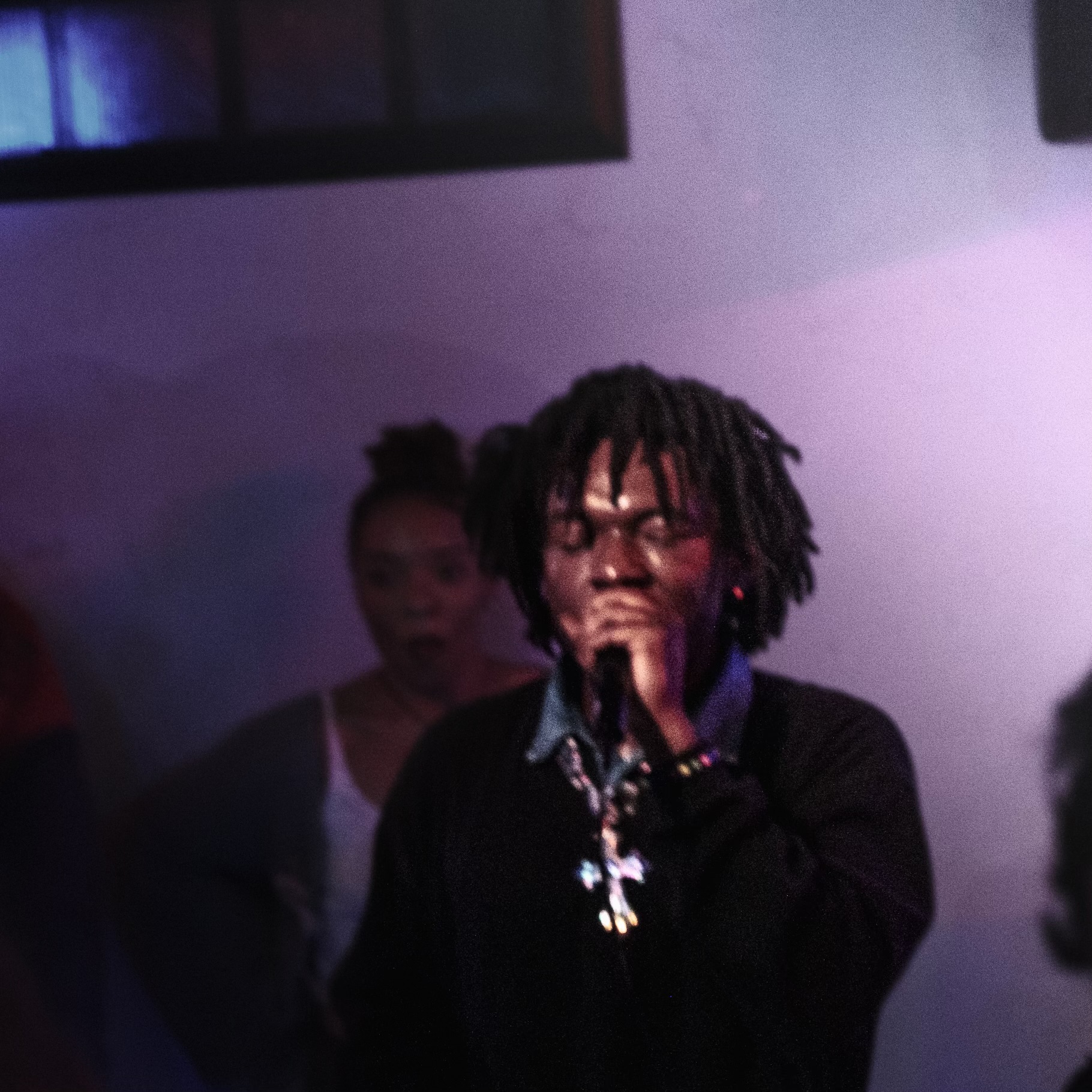 Person with dreadlocks singing into a microphone, wearing a dark jacket and jewelry, in a dimly lit room.