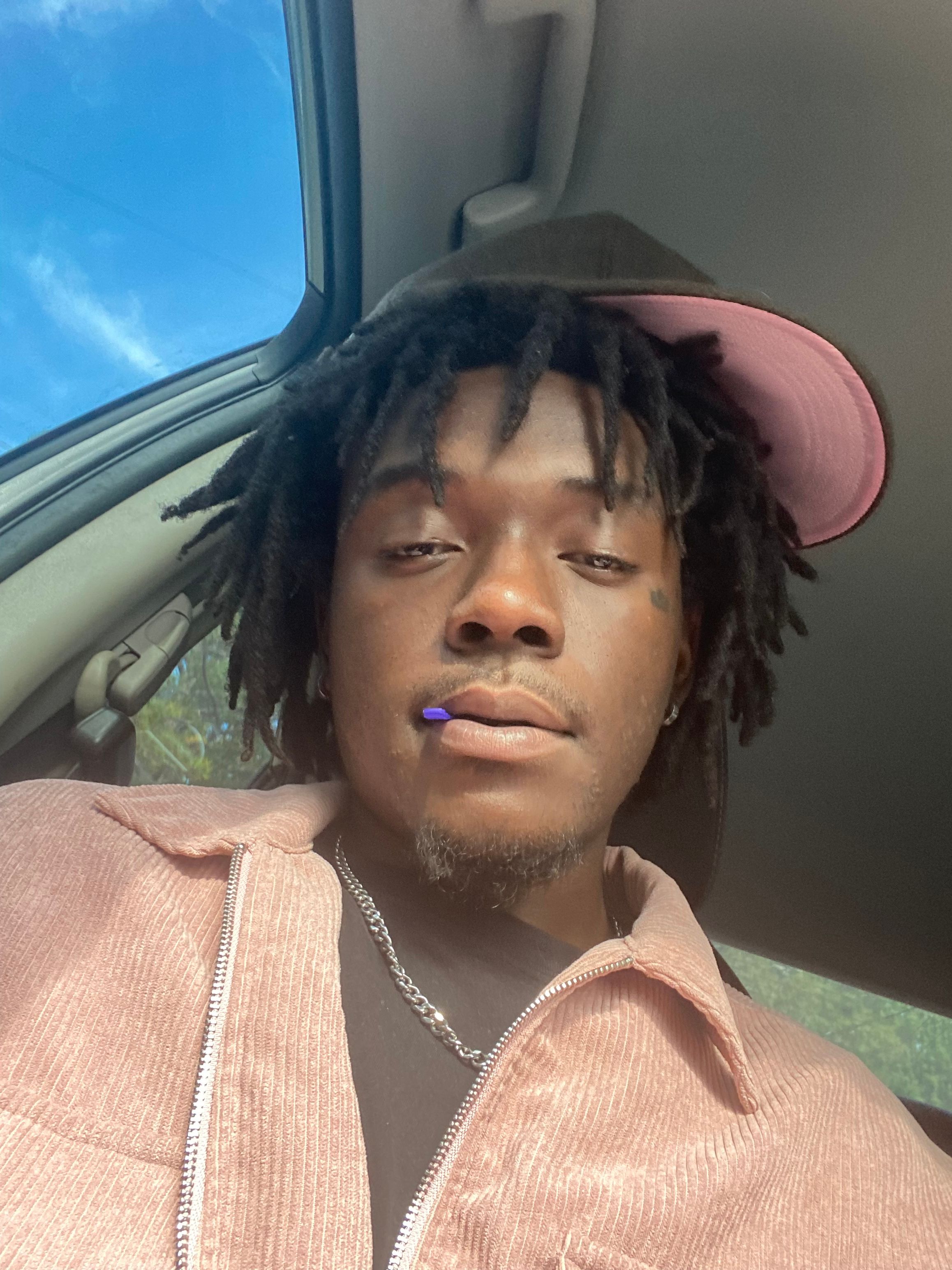 Young person with dreadlocks wearing a pink cap and beige jacket, sitting in a vehicle, looking at the camera.