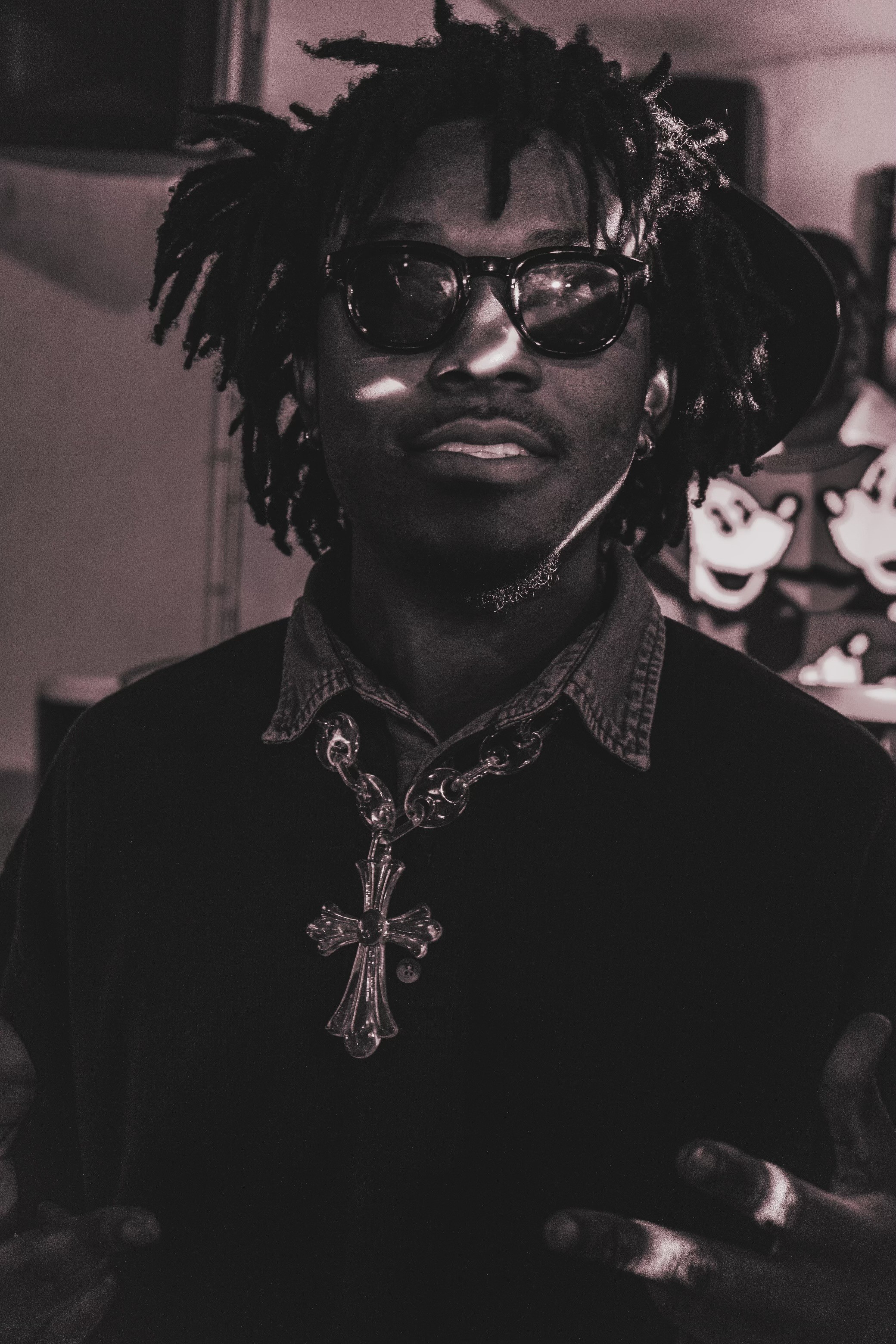 Person with dreadlocks, glasses, and a cross necklace, in a dark setting with cartoon character images in the background.