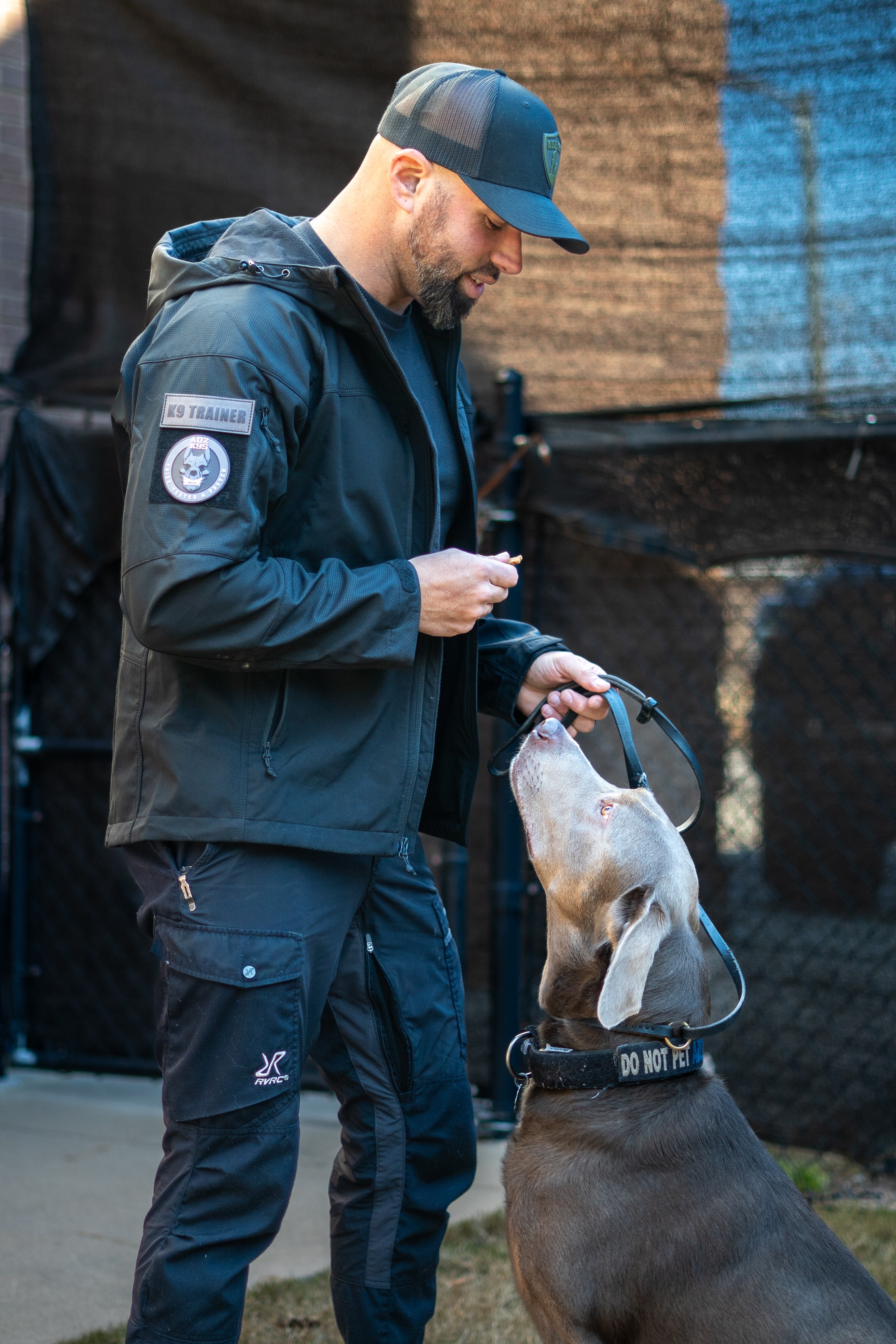 Man in dark jacket and cap smiling at dog holding a leash, outdoors, background with fence and tarp