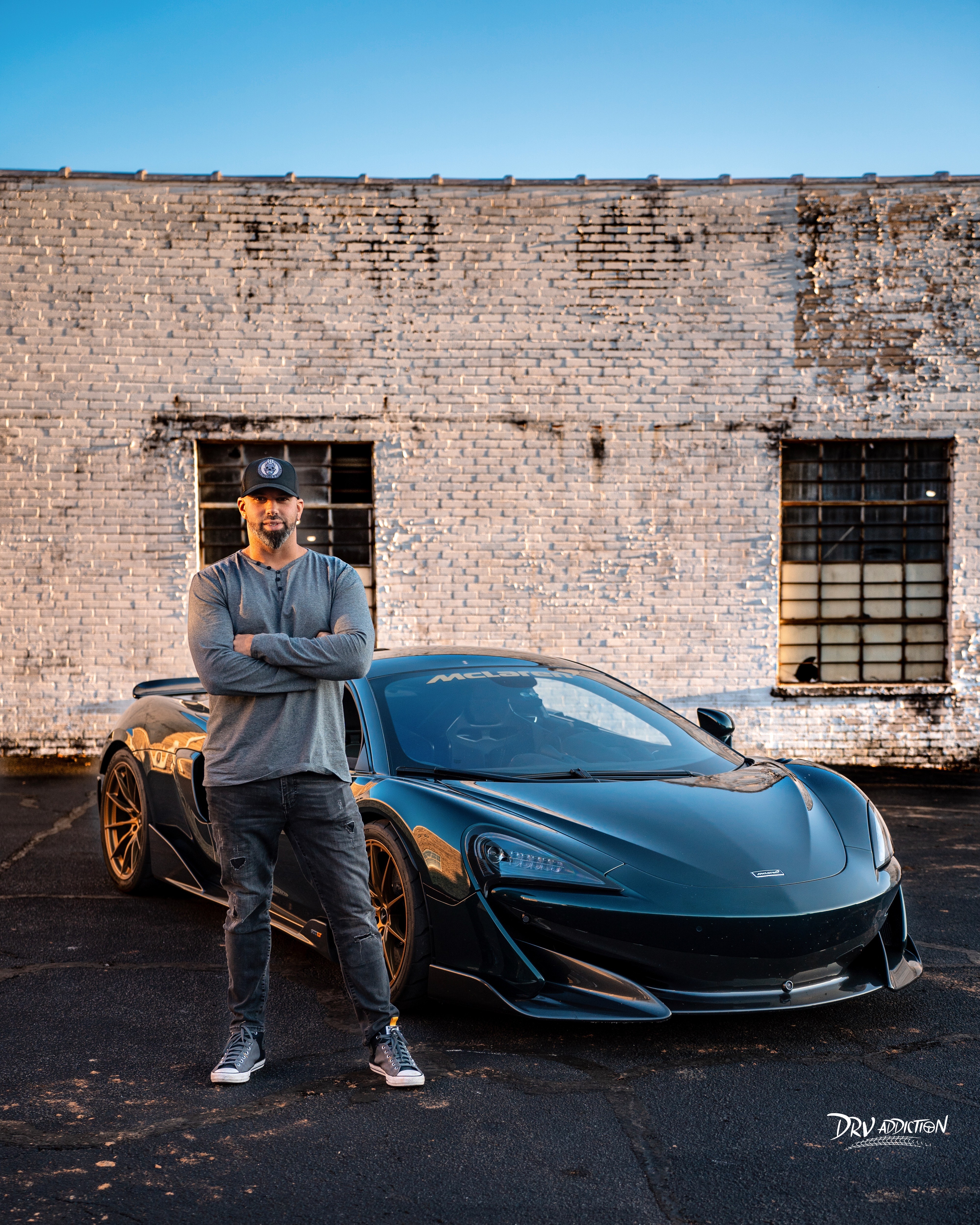 Man standing with arms crossed in front of a black sports car against a brick wall, clear blue sky above.