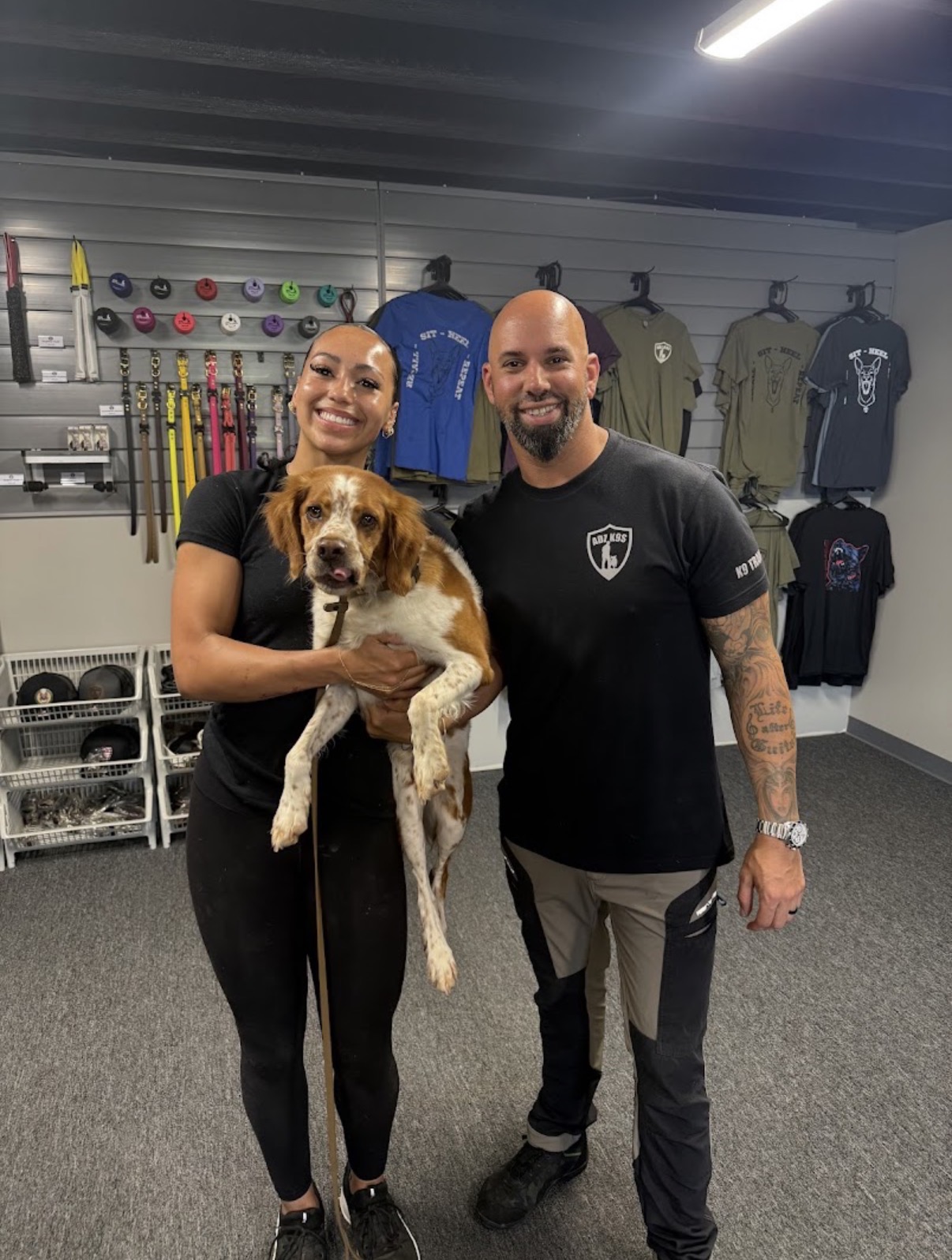 Two people and a dog in a gym, smiling, with fitness equipment and clothing in the background.