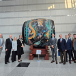 Group of people standing beside a large, decorated drum on a stand in a modern indoor space.