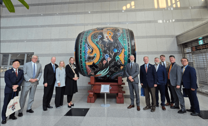 Group of people standing beside a large, decorated drum on a stand in a modern indoor space.