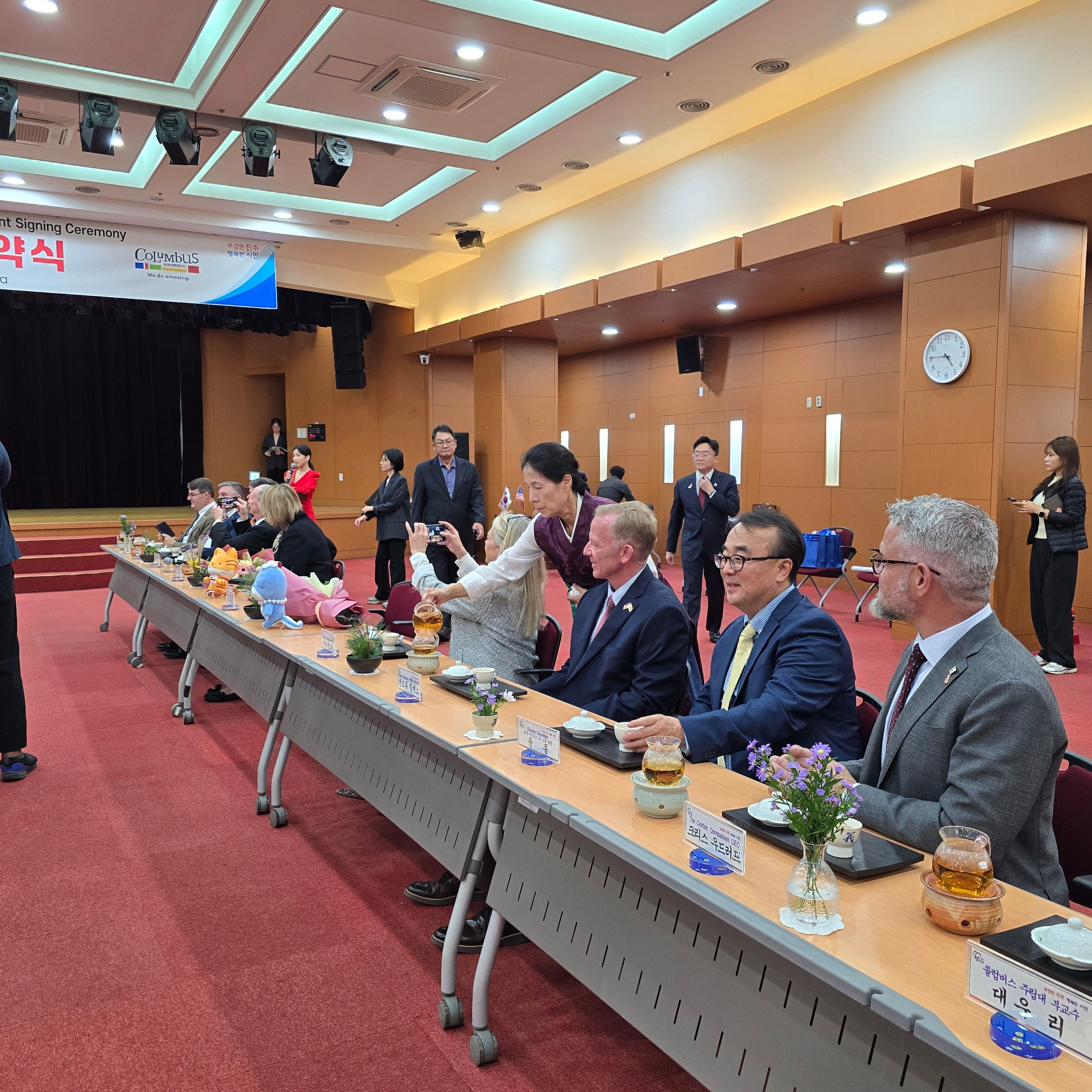 People gathered around a long table with food and drinks in a conference room, some standing and some seated, with a clock on the wall.