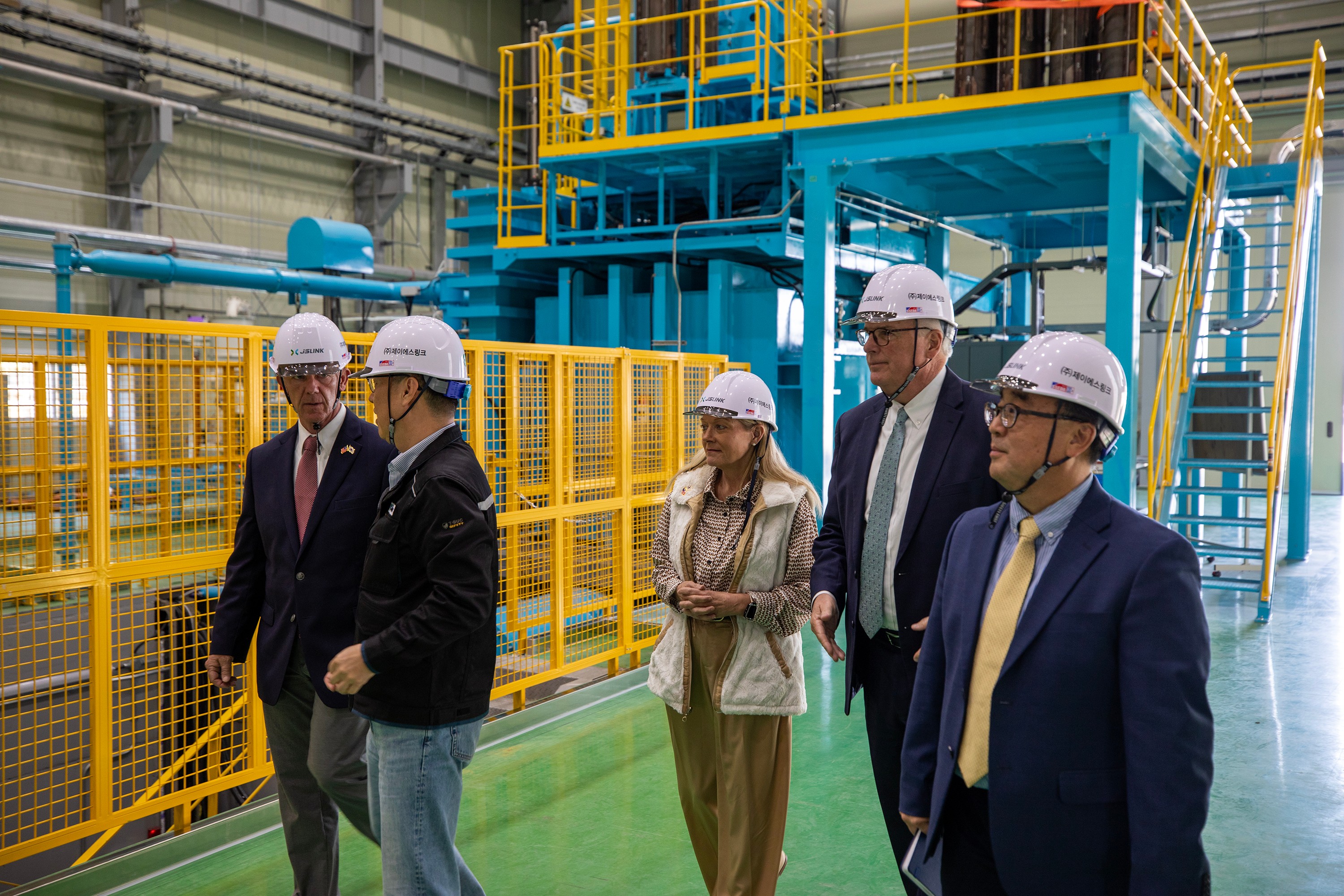 Group of five people wearing safety helmets walking inside an industrial facility with machinery and yellow safety barriers.