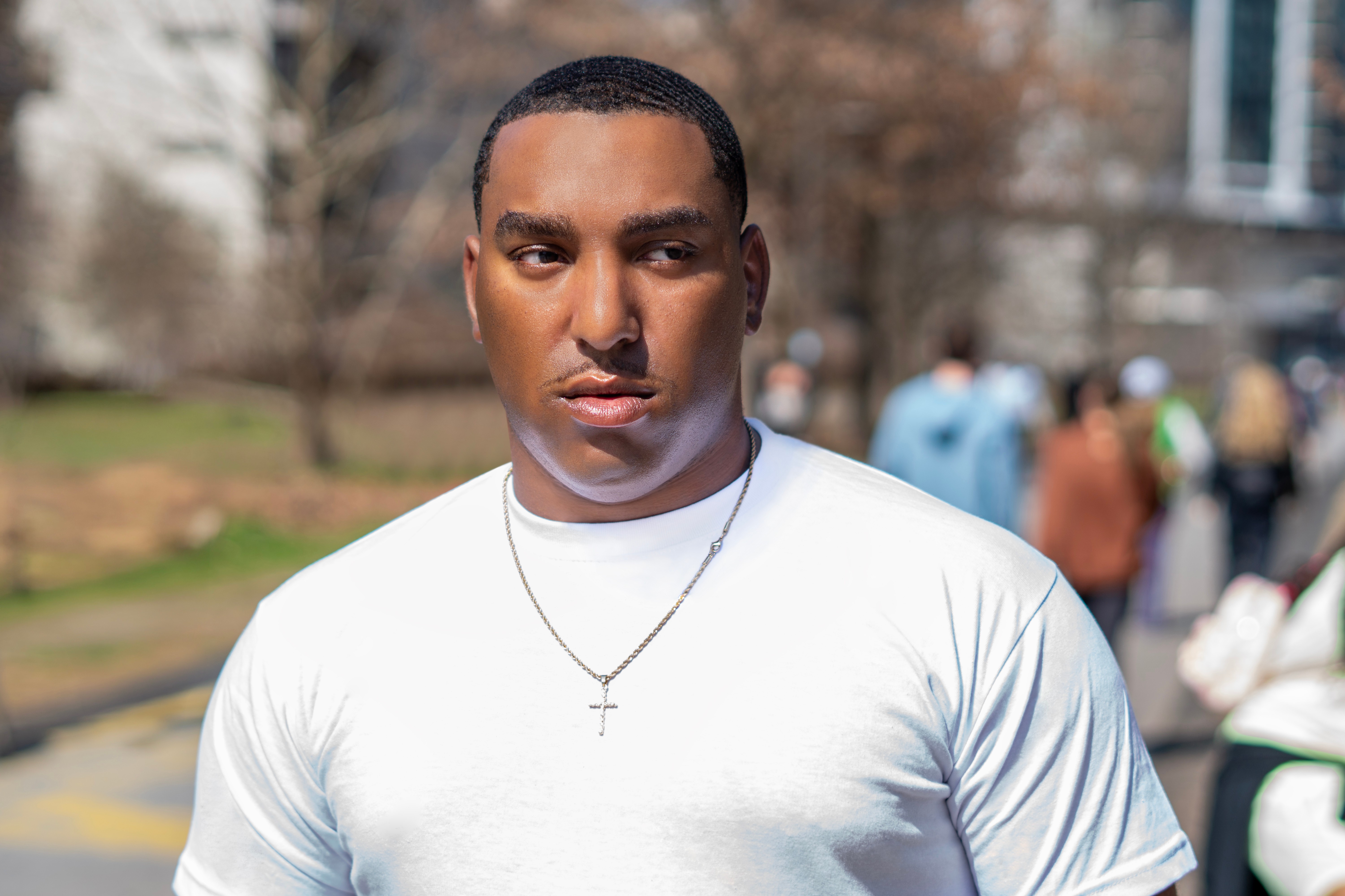 Young man with short hair wearing a white t-shirt and a cross necklace outdoors, with blurred people in background.