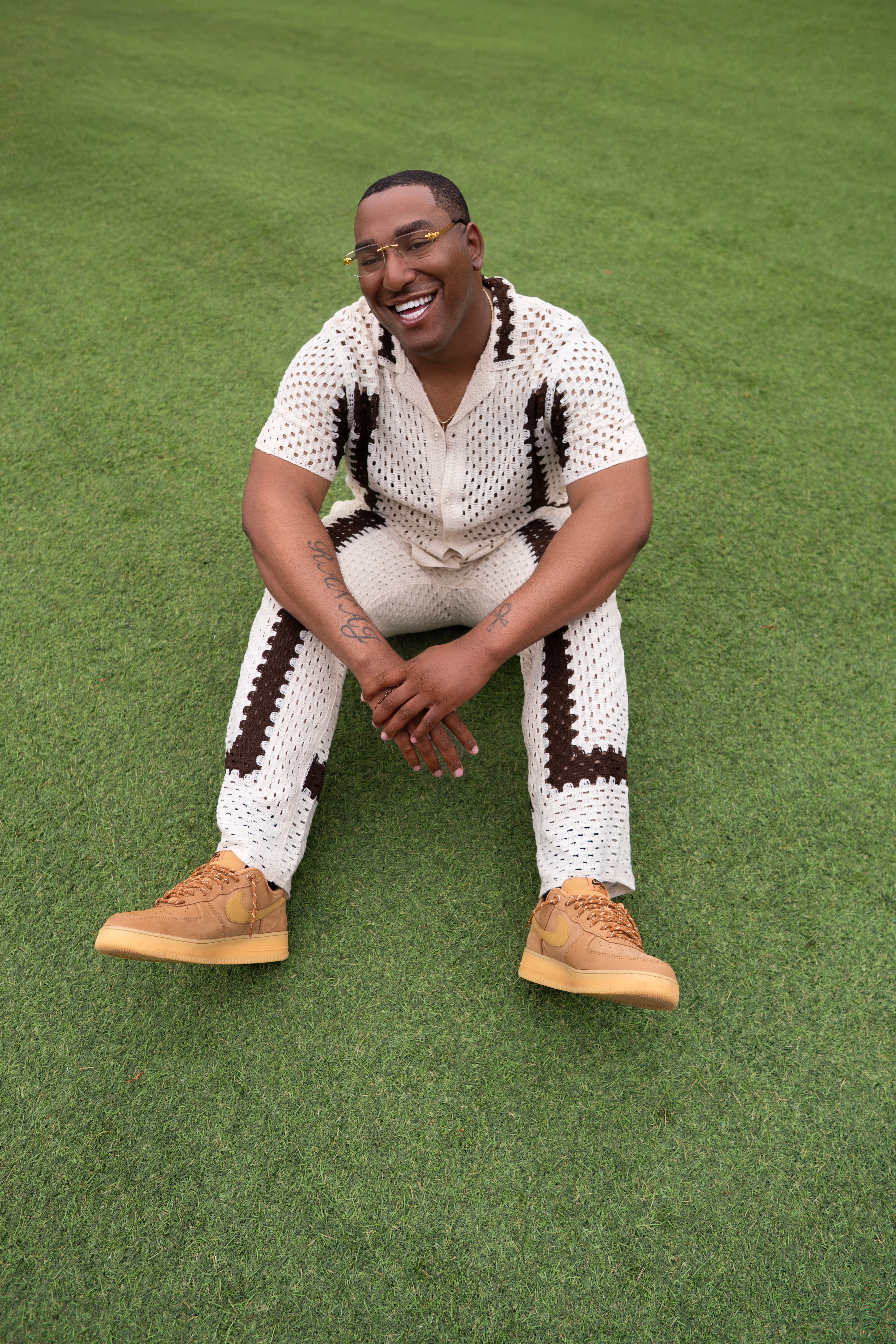 Person sitting on grass, smiling, wearing a white outfit with black patterns and tan shoes.