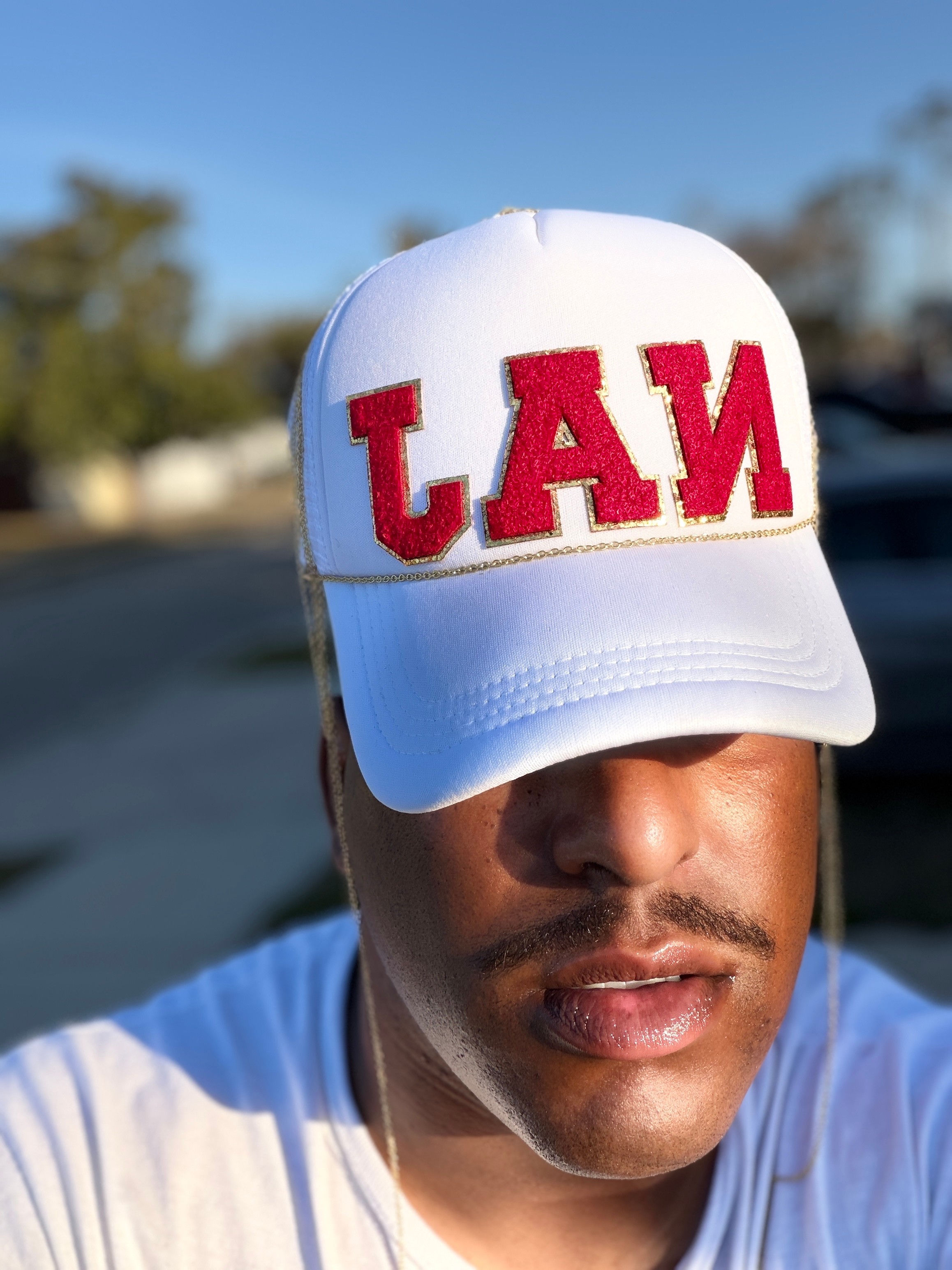 Person wearing a white cap with red embroidered text, outdoors with trees and blue sky in background.