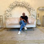 Person sitting on a cream-colored sofa with floral arch behind, gold mirror to the right, and decorative items around.