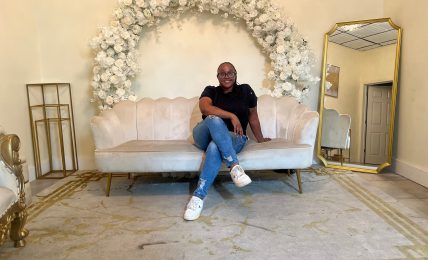 Person sitting on a cream-colored sofa with floral arch behind, gold mirror to the right, and decorative items around.