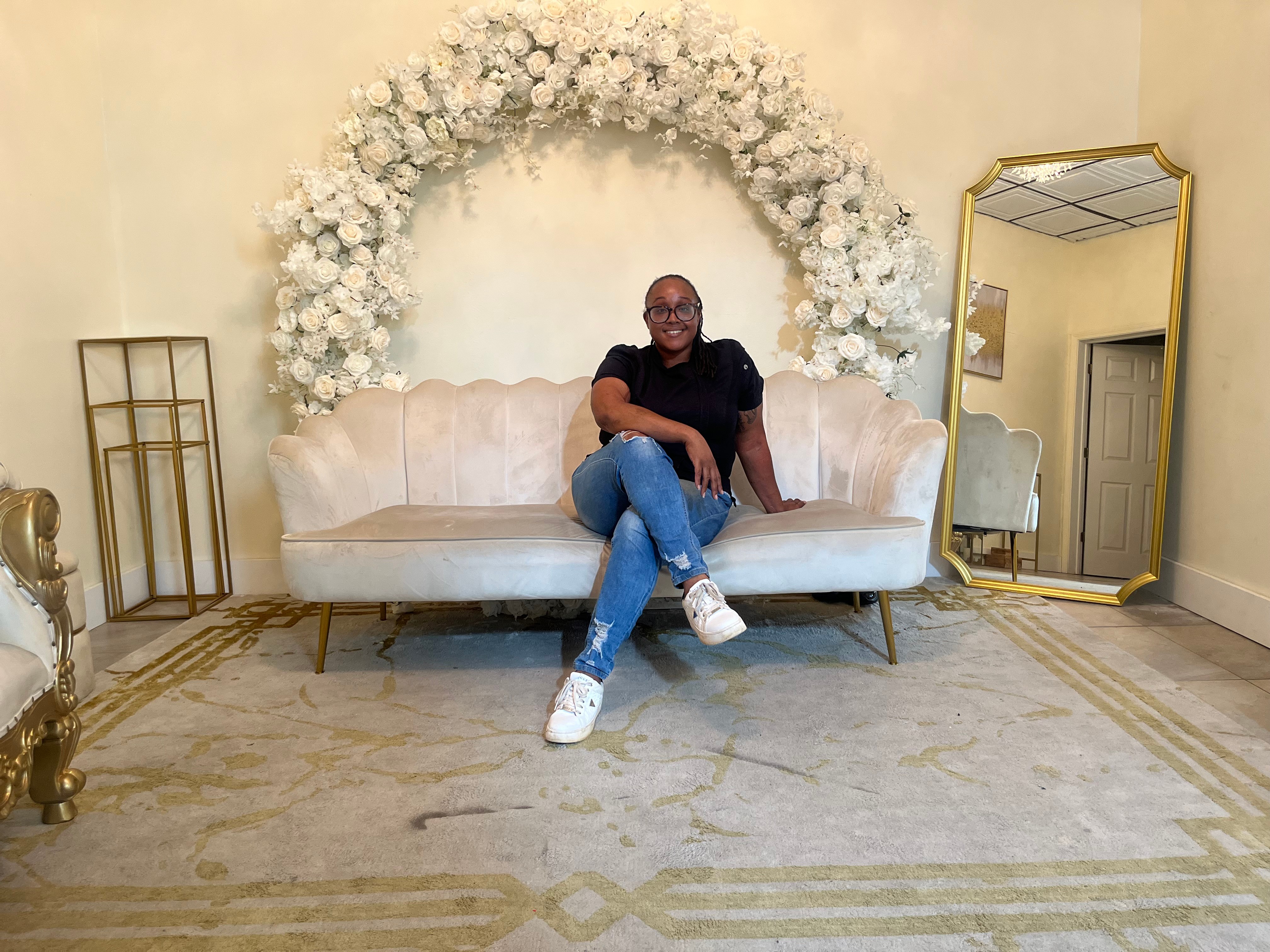 Person sitting on a cream-colored sofa with floral arch behind, gold mirror to the right, and decorative items around.