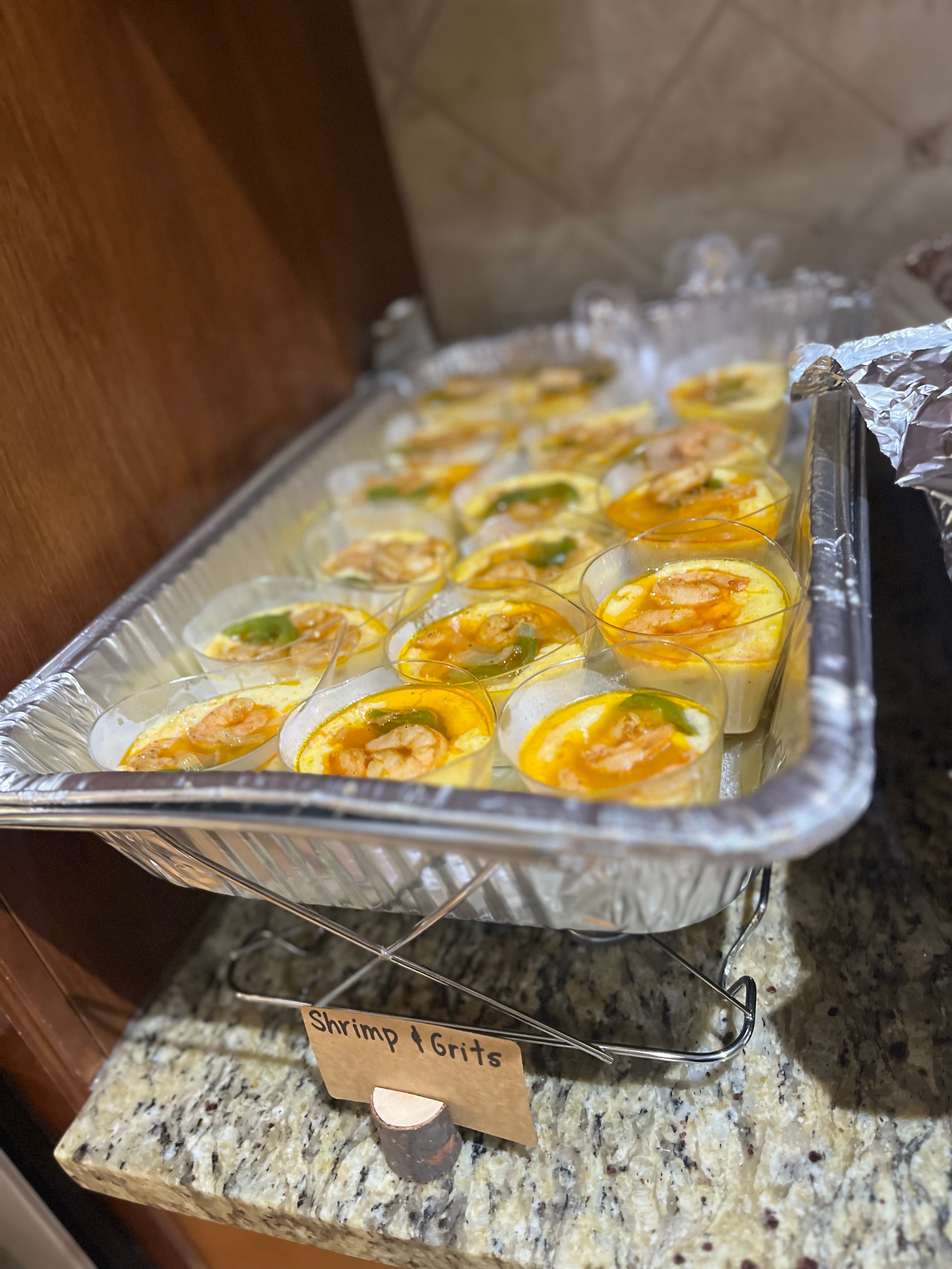 Tray of shrimp and guacamole appetizers on a countertop, with a handwritten label reading 'Shrimp & Grita', aluminum foil, and a wooden wall in the background.